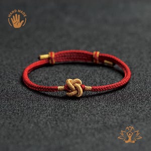 May include: A handmade red bracelet with a gold knot and gold accents. The bracelet is made of woven material and features a clasp closure. The words "HAND MADE" are printed on the upper left of the image.