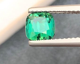 Puede incluir: Una vibrante piedra preciosa verde esmeralda sujeta con pinzas. La piedra de talla cojín muestra facetas que reflejan la luz, creando un efecto brillante. El color de la gema es un verde rico y saturado. Las pinzas son de color plateado.