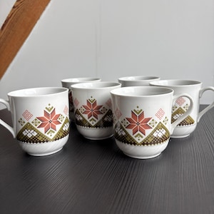 Könnte beinhalten: Ein Set aus sechs weißen Keramikbechern mit einem dekorativen Muster. Das Muster zeigt ein zentrales rot-oranges Sternmuster, umrahmt von grünen und braunen geometrischen Formen. Jeder Becher hat einen weißen Henkel.
