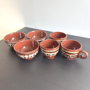Pode incluir: Um conjunto de seis taças e chávenas de cerâmica com interior em terracota brilhante e uma faixa exterior decorativa com um padrão de esmalte escorrido em tons de creme, castanho escuro e bege. O conjunto inclui taças e uma chávena com asa.