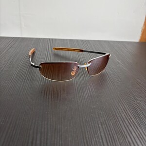May include: Rimless sunglasses with brown tinted lenses. The frames are silver with black accents and brown temples. The sunglasses are resting on a dark brown wooden surface.