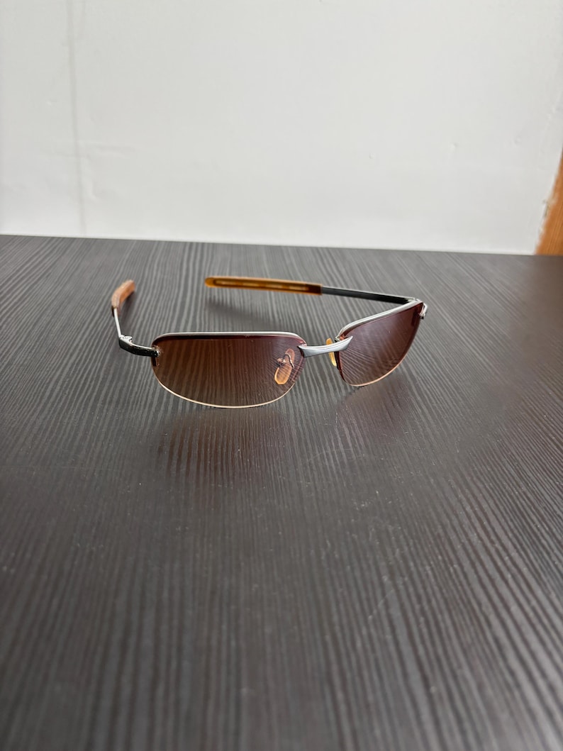 May include: Rimless sunglasses with brown tinted lenses. The frames are silver with black accents and brown temples. The sunglasses are resting on a dark brown wooden surface.