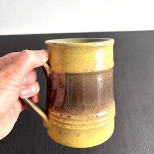 Può includere: Una tazza in ceramica con corpo cilindrico e smalto bicolore. Le sezioni superiore e inferiore sono di un giallo senape brillante, mentre la fascia centrale è di un marrone scuro maculato. Un manico è fissato sul lato della tazza.