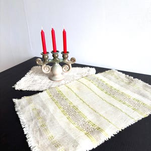 Könnte beinhalten: Ein Kerzenständer aus Keramik mit drei Armen und leuchtend roten Stabkerzen, begleitet von zwei rechteckigen Web-Platzsets. Die Platzsets haben eine hellbeige Basis mit vertikalen Streifen in gedecktem Grün und Grau sowie ausgefransten Rändern.