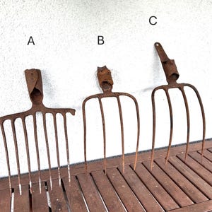 May include: Three rusted metal garden forks labeled A, B, and C. Fork A has seven tines, while forks B and C each feature four tines. Each tool includes a cylindrical socket at the top for handle attachment, showing significant oxidation and wear.