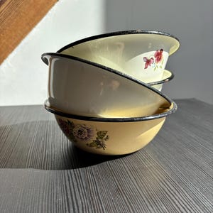 Op de afbeelding: Drie vintage emaille kommen op elkaar gestapeld. Ze zijn crèmekleurig met zwarte randen en bloemmotieven in roze en paars. De kommen staan op een donkergrijze ondergrond. Elke kom heeft een diameter van ongeveer 20 cm.