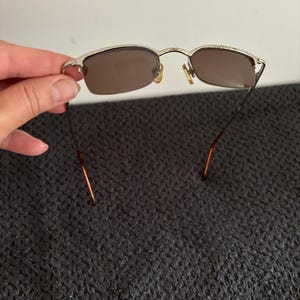 May include: Silver-framed sunglasses with brown lenses. The rectangular lenses are set in a silver frame with a thin bridge and temples. The sunglasses are held against a dark gray textured background.