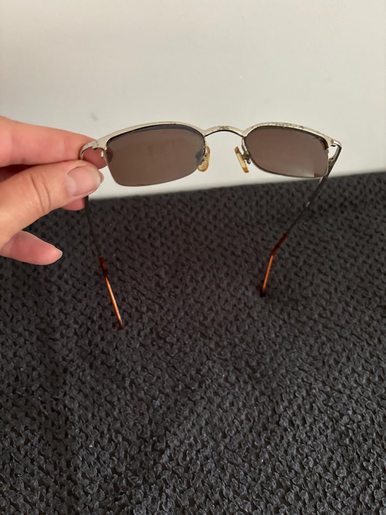May include: Silver-framed sunglasses with brown lenses. The rectangular lenses are set in a silver frame with a thin bridge and temples. The sunglasses are held against a dark gray textured background.