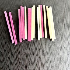 Op de afbeelding: Een verzameling van negentien dunne, rechthoekige plastic staafjes die in een losse groep zijn gerangschikt. De staafjes zijn gekleurd in tinten felroze, lichtgeel en gebroken wit, elk met een glad, plat oppervlak en rechte randen.
