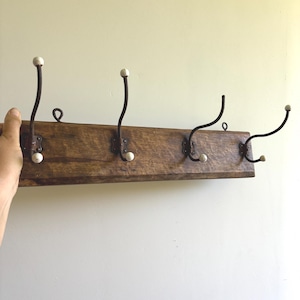 May include: A rectangular wooden wall rack featuring four dark metal hooks. Each hook has a curved design with small white ceramic knobs at the top and bottom ends. The wood has a natural brown finish with visible grain and texture throughout the surface.