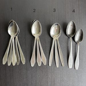 Op de afbeelding: Vier sets vintage zilveren lepels zijn gerangschikt op een donkere ondergrond. Elke set heeft een ander ontwerp op het handvat. De lepels zijn gemaakt van een glanzend, reflecterend metaal, waarschijnlijk zilver.