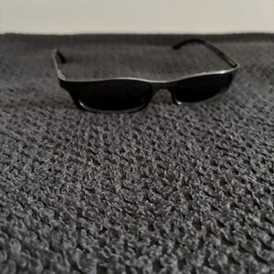 May include: Black sunglasses with a silver trim on the frame. The sunglasses are rectangular and have dark lenses. The arms of the sunglasses are black. The sunglasses are resting on a textured, dark gray surface.