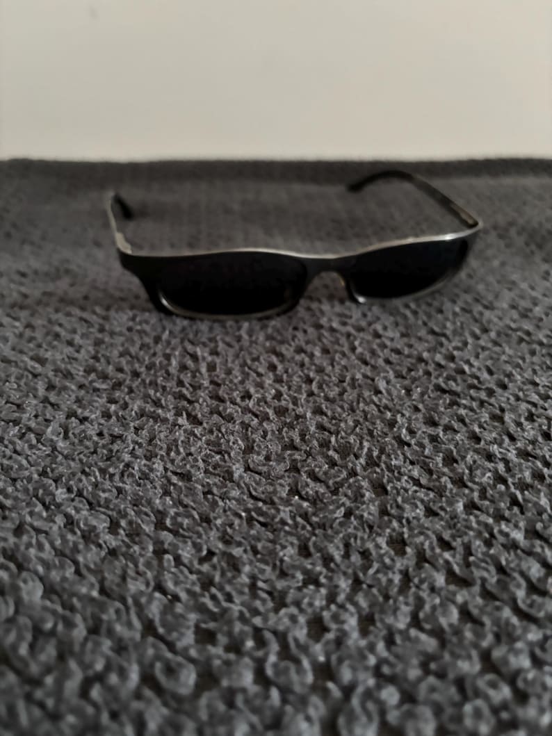 May include: Black sunglasses with a silver trim on the frame. The sunglasses are rectangular and have dark lenses. The arms of the sunglasses are black. The sunglasses are resting on a textured, dark gray surface.