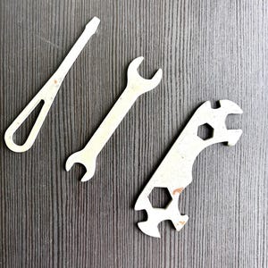 Può includere: Un set di tre utensili in metallo color argento composto da un cacciavite a testa piatta con manico aperto, una chiave fissa a doppia estremità e una chiave curva multifunzionale con due aperture esagonali e bordi dentellati per la presa.