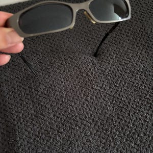 May include: Silver-framed sunglasses with dark lenses. The sunglasses have a rectangular shape and a thin frame. The temples are black and extend from the frame. The sunglasses are held by a hand.