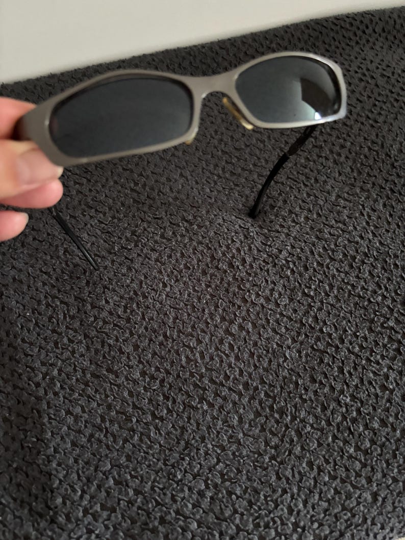 May include: Silver-framed sunglasses with dark lenses. The sunglasses have a rectangular shape and a thin frame. The temples are black and extend from the frame. The sunglasses are held by a hand.