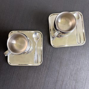 Puede incluir: Dos tazas de espresso a juego de acero inoxidable con asas, cada una sobre una bandeja cuadrada con una cuchara pequeña. Las tazas y las bandejas tienen una superficie brillante y reflectante. El fondo es una superficie oscura y texturizada.