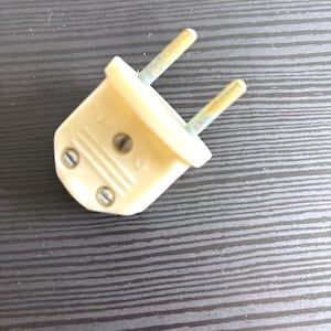 Può includere: Una spina elettrica vintage a due poli di colore bianco sporco, caratterizzata da un corpo in plastica stampata con due viti metalliche visibili sulla parte anteriore e due spinotti metallici che sporgono, con segni di usura e scolorimento.