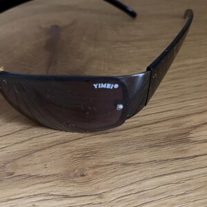 May include: Black sunglasses with dark tinted lenses. The frame is black and has the text "YIMBIO" printed on the side. The sunglasses are resting on a wooden surface.