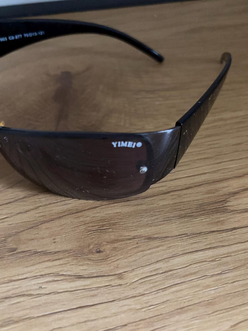 May include: Black sunglasses with dark tinted lenses. The frame is black and has the text "YIMBIO" printed on the side. The sunglasses are resting on a wooden surface.