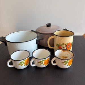 May include: A selection of vintage enamel kitchenware. The image shows a large white pot, a grey lidded pot, a yellow mug with floral detail, and three white mugs with orange rose designs. The items are arranged on a dark surface.