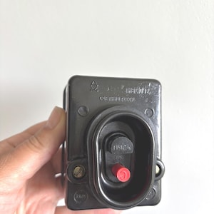 Peut inclure: Un interrupteur de démarrage électrique rectangulaire noir doté d'un centre encastré avec un petit bouton rouge et un bouton noir portant l'inscription cyrillique "ПУСК". Le boîtier comporte du texte cyrillique moulé et un petit logo.