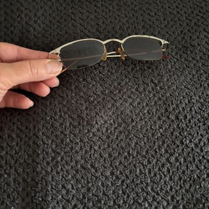 May include: Rimless sunglasses with a silver metal frame and dark lenses. The glasses have a rectangular shape and are held by a hand against a dark background. The temples are thin and extend to the ear pieces.