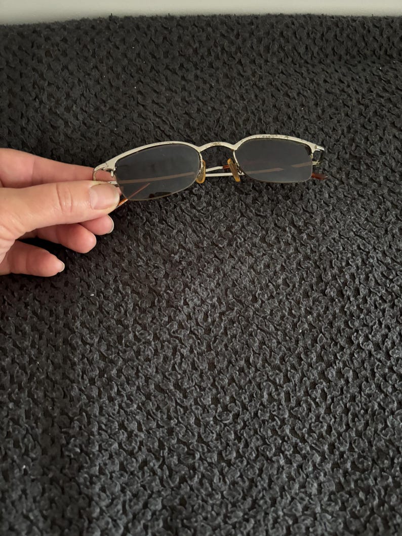 May include: Rimless sunglasses with a silver metal frame and dark lenses. The glasses have a rectangular shape and are held by a hand against a dark background. The temples are thin and extend to the ear pieces.