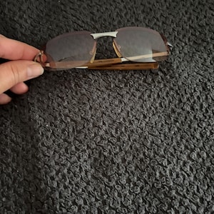 May include: Rimless sunglasses with brown tinted lenses and a silver bridge. The frames have a tortoise shell pattern. The sunglasses are folded.