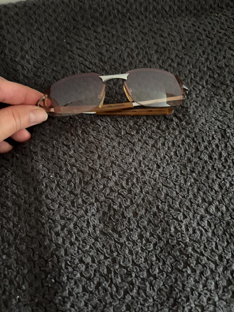 May include: Rimless sunglasses with brown tinted lenses and a silver bridge. The frames have a tortoise shell pattern. The sunglasses are folded.