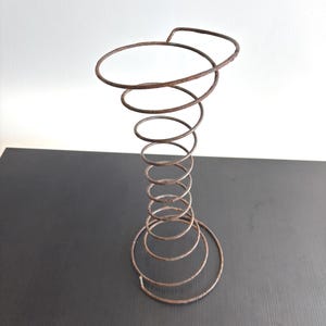 Può includere: Una molla di metallo arrugginita, a spirale, con una parte superiore e inferiore circolari. La molla è di colore marrone scuro e si trova su una superficie scura contro uno sfondo bianco. Un oggetto vintage.