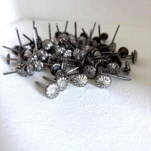 May include: A collection of silver-toned metal upholstery tacks featuring a circular head with a raised, radiating sunburst or floral pattern and a sharp, pointed nail extending from the center of the underside of each individual fastener.