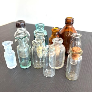 May include: A collection of eleven small glass bottles in various shapes and sizes, including clear, amber, and light blue glass. Some bottles feature narrow necks, one has a dark brown screw cap, and one contains a small cork stopper.