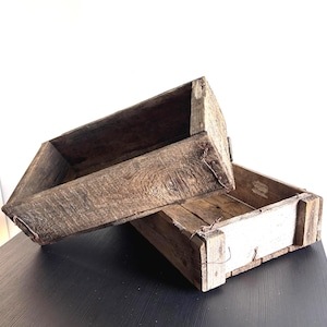 Puede incluir: Dos cajas de madera rectangulares con un acabado marrón oscuro desgastado. Las cajas presentan vetas de madera visibles, texturas rugosas y cierres de alambre metálico expuestos en las esquinas. Una caja está colocada en ángulo sobre la otra.