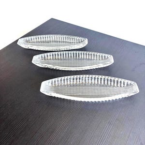 Peut inclure: Trois plats en verre transparent de forme ovale allongée. Chaque plat présente un motif nervuré texturé le long du bord extérieur et une série de lignes horizontales parallèles gravées sur la base plate et centrale de la surface en verre.