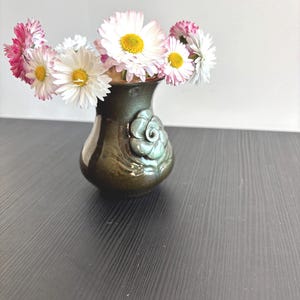 Può includere: Un piccolo vaso in ceramica verde scuro con finitura lucida e un rilievo floreale scolpito sulla parte anteriore. Il vaso è riempito con un bouquet di fiori simili a margherite bianche e rosa con centri gialli vivaci e contrastanti.