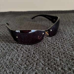 May include: Black sunglasses with a curved, rimless design. The lenses are a dark, reflective color, and the frames are black. Small gold-colored accents are visible near the hinges. The sunglasses are resting on a textured, gray surface.