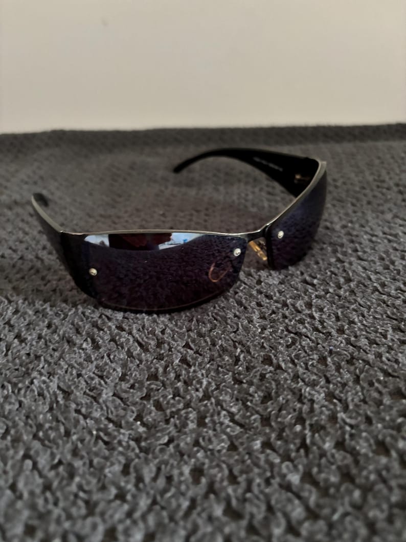 May include: Black sunglasses with a curved, rimless design. The lenses are a dark, reflective color, and the frames are black. Small gold-colored accents are visible near the hinges. The sunglasses are resting on a textured, gray surface.