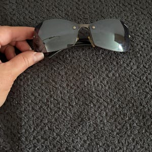 May include: Rimless sunglasses with dark tinted lenses and a silver-tone metal frame. The sunglasses have a rectangular shape and are held by a hand against a dark textured background.