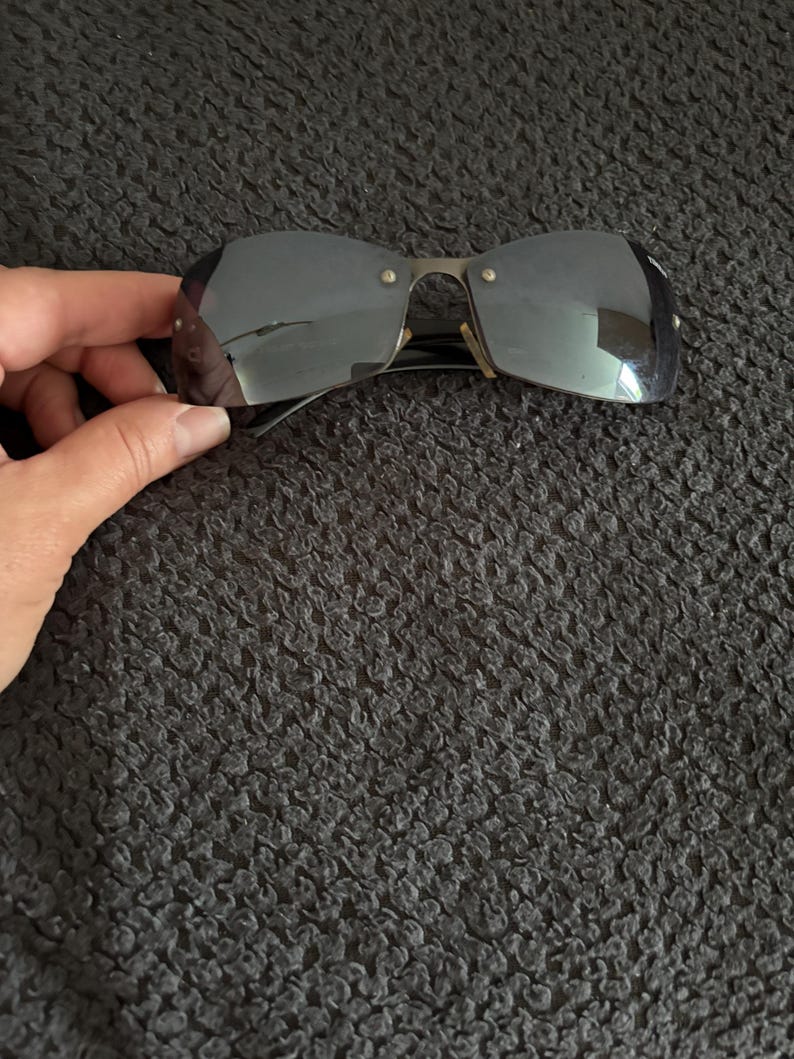 May include: Rimless sunglasses with dark tinted lenses and a silver-tone metal frame. The sunglasses have a rectangular shape and are held by a hand against a dark textured background.