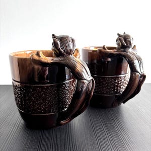 Può includere: Due tazze in ceramica marrone scuro con un manico scolpito a forma di scoiattolo. Le tazze presentano un bordo superiore liscio e una fascia centrale testurizzata e granulosa. La figura dello scoiattolo è modellata come se si arrampicasse.