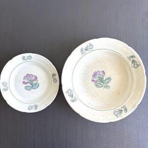 May include: A small plate and a larger bowl, both off-white with scalloped edges. Each piece features a central purple and green floral illustration and smaller green floral motifs spaced evenly around the rim of the ceramic dinnerware set.