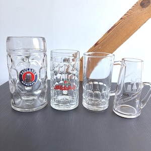 Op de afbeelding: Vier heldere glazen bierpullen van verschillende maten en ontwerpen. Eén pul heeft het "Paulaner Munchen" logo, een andere heeft "Alda" in rood, en een derde heeft een "100" afbeelding. De pullen staan op een donkergrijze ondergrond.