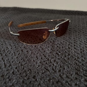 May include: Rimless sunglasses with brown tinted lenses and silver metal frames. The temples are a light brown wood grain. The sunglasses are resting on a dark gray textured surface.