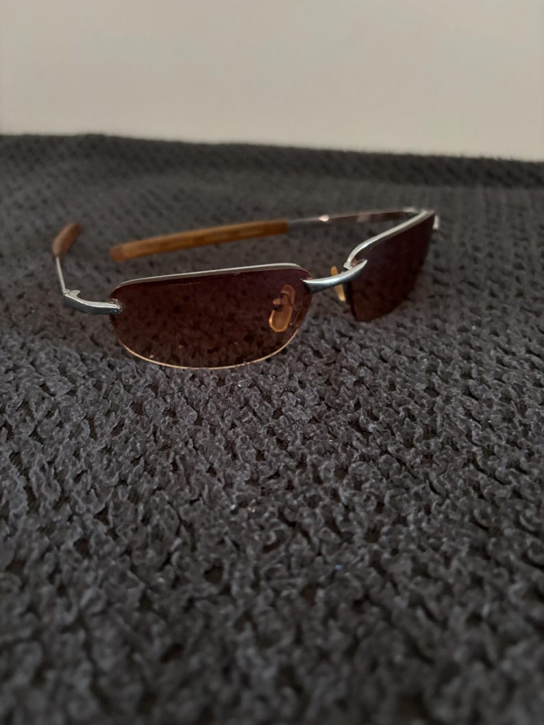 May include: Rimless sunglasses with brown tinted lenses and silver metal frames. The temples are a light brown wood grain. The sunglasses are resting on a dark gray textured surface.