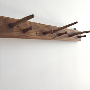 Könnte beinhalten: Eine rechteckige Wandgarderobe aus Holz mit einer Reihe von hervorstehenden Haken zum Aufhängen von Gegenständen. Die Leiste besteht aus naturfarbenem braunem Holz mit sichtbarer Maserung und mehreren konischen Stiften, die nach außen ragen.