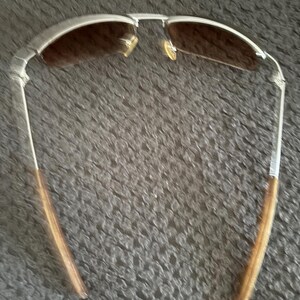 May include: Silver-framed sunglasses with brown tinted lenses. The sunglasses have a classic aviator style with a double bridge and slender temples. The temples are a light brown color. The sunglasses are designed to protect the eyes from the sun.