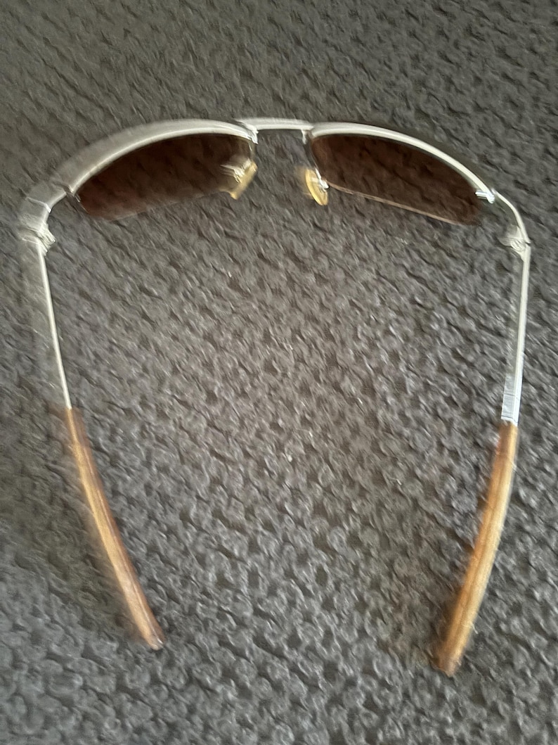 May include: Silver-framed sunglasses with brown tinted lenses. The sunglasses have a classic aviator style with a double bridge and slender temples. The temples are a light brown color. The sunglasses are designed to protect the eyes from the sun.