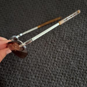 May include: Sunglasses with a silver metal frame and brown tinted lenses. The temples are silver with a brown accent near the ear. The sunglasses are angled, showing the shape of the lenses and the design of the frame.