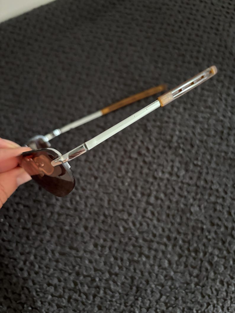 May include: Sunglasses with a silver metal frame and brown tinted lenses. The temples are silver with a brown accent near the ear. The sunglasses are angled, showing the shape of the lenses and the design of the frame.